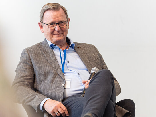 Bodo Ramelow sitzt während einer Veranstaltung auf der Bühne und lächelt freundlich ins Publikum. 