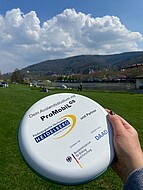 Das Bild zeigt eine Frisbee, auf der insbesondere der Projekttitel und die Logos der beteiligten Hochschule zu sehen sind. Im Hintergrund ist unscharf der Neckar und der Königstuhl zu sehen. 