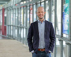 Die Portraitaufnahme zeigt Martin Giese. Er steht in einem Gang mit großen Fensterscheiben. Den Hintergrund sieht man lediglich verschwommen. Copyright: privat