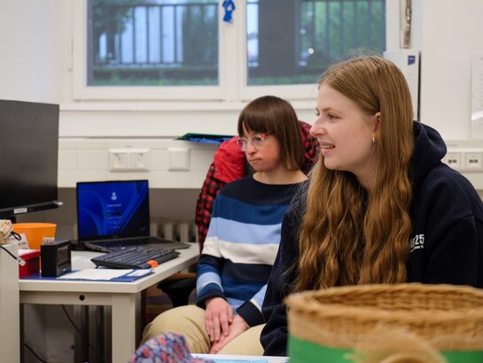 Anne Genzwürker sitzt an einem Schreibtisch, im Hintergund ist Bildungsfachkraft Susann Bensch