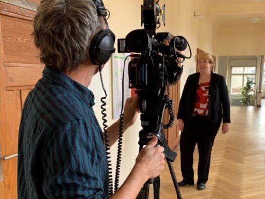 Das Bild zeigt im Vordergrund eine Person mit einer großen Videokamera. Und im Hintergrund ist Frau Terfloth zu erkennen.