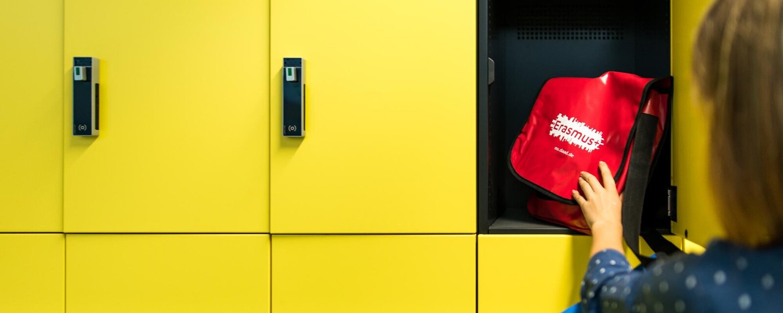 gelber Spind in den eine Person einen roten Rucksack legt