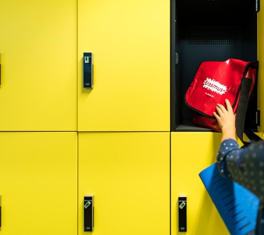 gelber Spind in den eine Person einen roten Rucksack legt