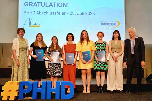 Zu sehen sind Alumni, die auf einer Bühne stehen und ihre Bestehensbriefe in PHHD-Mappen hochhalten. Gemeinsam mit Rektorin Vach & Prorektor Dammer (a.D.), welche ihnen die Mappen überreicht haben.