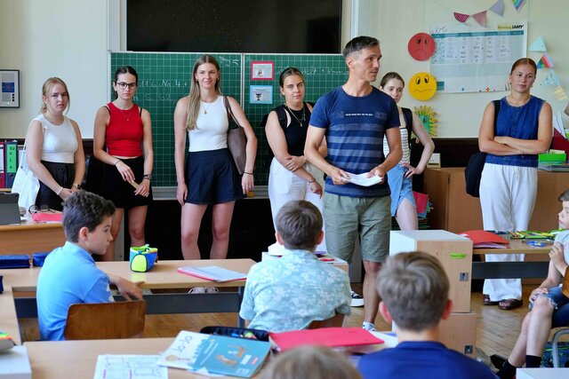 Auch im Klassenzimmer findet digitale Lehre statt: Zu sehen sind Studierende vor einer Tafel, sowie ein Dozent. Kinder sitzen im Klassenzimmer und schauen zur Tafel. 