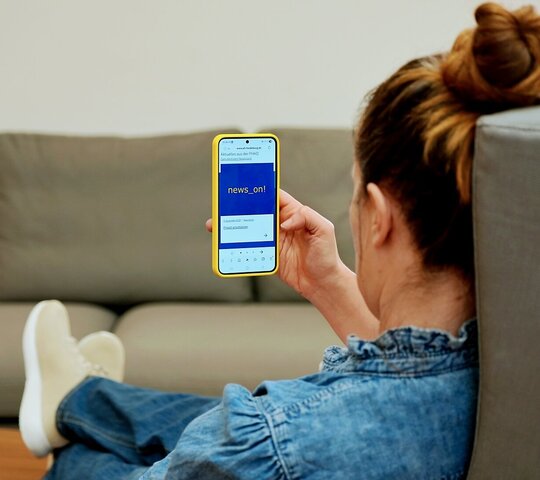 Zu sehen ist eine Frau in einem Sessel, die auf dem Smartphone den Newsletter liest