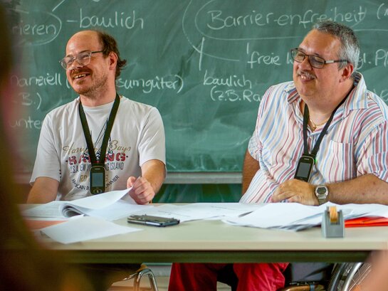 Das Bild zeigt die Bildungsfachkräfte Thilo Krahnke und Helmuth Pflantzer, wie sie ein Bildungsangebot ausbringen. Beide sitzen vor einer Tafel, auf der unter anderem die Worte "Barrierefreihet" zu lesen sind.