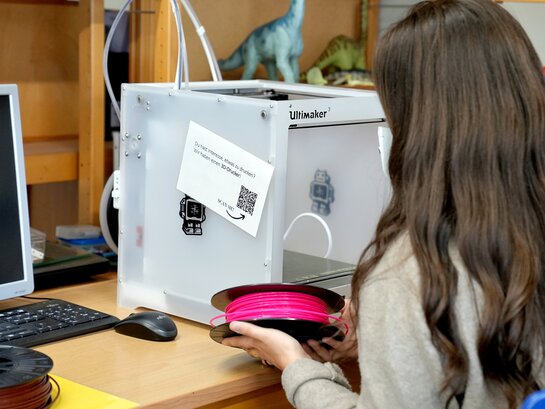 Studentin sitzt vor 3 Drucker und hält Plastikrolle in der Hand