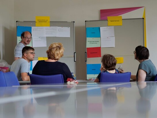 Das Bild zeigt im Hintergund mehrere Personen, die diskutieren. Eine Person steht vor einer Pinnwand, an der unterschiedlich farbige Zettel angepinnt sind. Vier Personen sitzen der Pinnwand gegenüber.