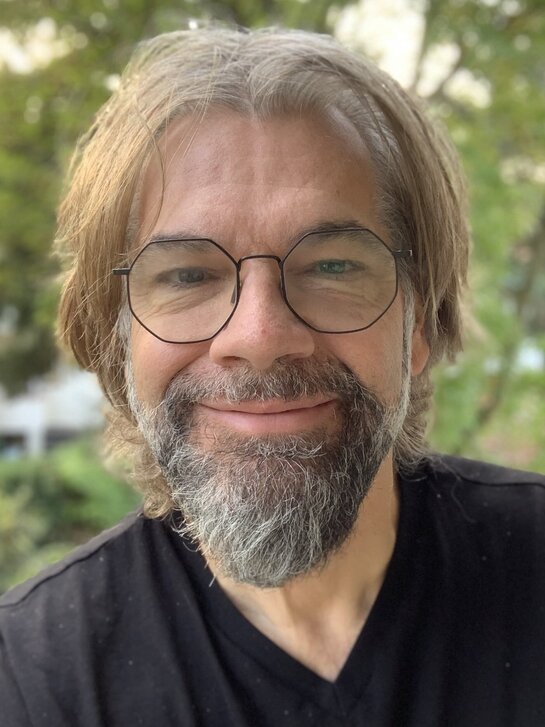 Das Bild zeigt Markus Fertig. Er trägt eine Brille, hat einen Bart und lächelt in die Kamera.