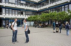 Das Bild zeigt den Neubau der Pädagogischen Hochschule Heidelberg. Vor diesem sind zahlreiche Studierende in Interaktion miteinander zu sehen.