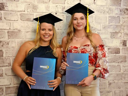 Zu sehen sind zwei Alumnae mit Graduierungshut in der Fotobox, wie sie ihre PHHD-Mappen hochhalten.