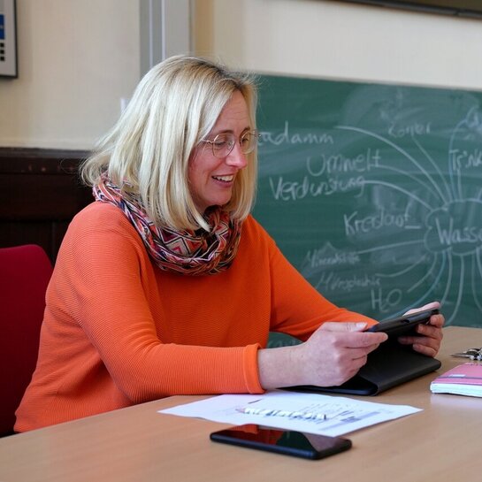 Eine Frau sitz in einem Klassenzimmer und schaut auf ein Tablet.