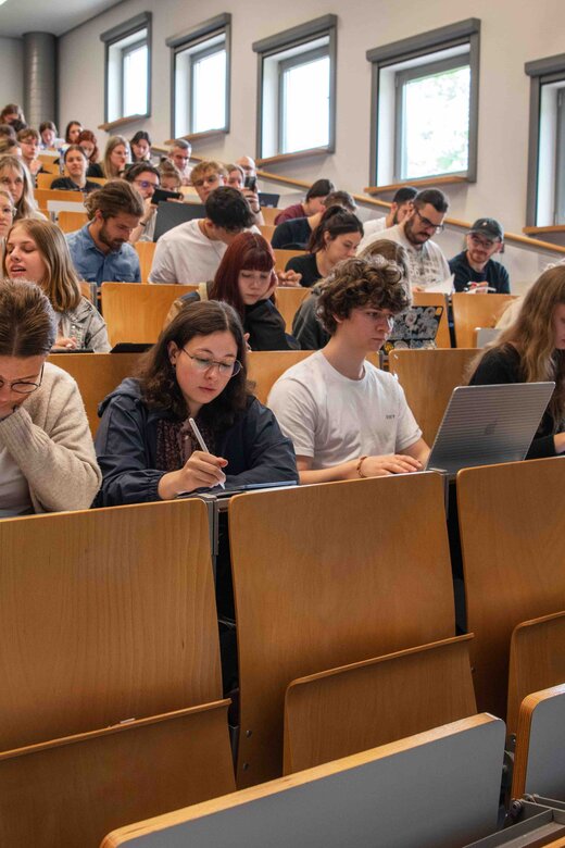 Ein Dozent steht vor einem vollen Hörsaal. Er beugt sich freundlich in Richtung der Studierenden. Das Bild wurde im Hörsaalgebäude der Hochschule aufgenommen.