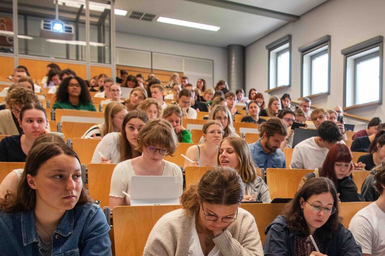Ein Dozent steht vor einem vollen Hörsaal. Er beugt sich freundlich in Richtung der Studierenden. Das Bild wurde im Hörsaalgebäude der Hochschule aufgenommen.