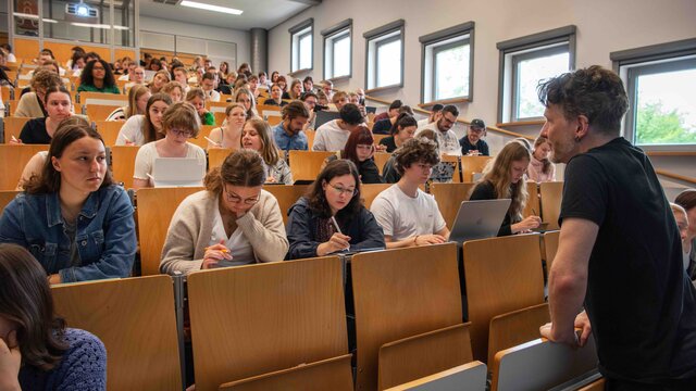 Ein Dozent steht vor einem vollen Hörsaal. Er beugt sich freundlich in Richtung der Studierenden. Das Bild wurde im Hörsaalgebäude der Hochschule aufgenommen.