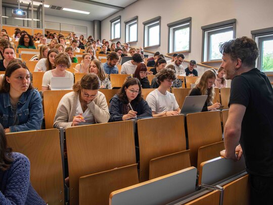 Ein Dozent steht vor einem vollen Hörsaal. Er beugt sich freundlich in Richtung der Studierenden. Das Bild wurde im Hörsaalgebäude der Hochschule aufgenommen.