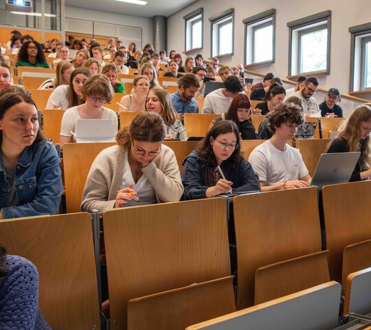 Ein Dozent steht vor einem vollen Hörsaal. Er beugt sich freundlich in Richtung der Studierenden. Das Bild wurde im Hörsaalgebäude der Hochschule aufgenommen.