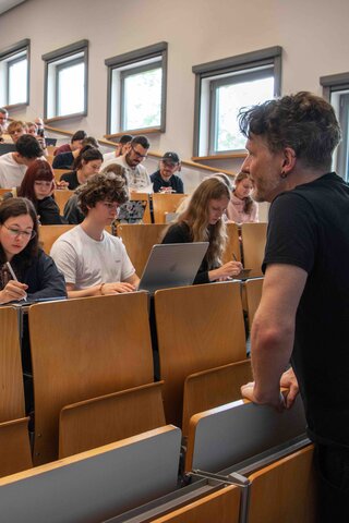 Ein Dozent steht vor einem vollen Hörsaal. Er beugt sich freundlich in Richtung der Studierenden. Das Bild wurde im Hörsaalgebäude der Hochschule aufgenommen.