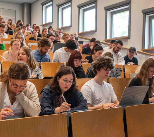 Ein Dozent steht vor einem vollen Hörsaal. Er beugt sich freundlich in Richtung der Studierenden. Das Bild wurde im Hörsaalgebäude der Hochschule aufgenommen.