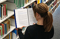 Auf dem Bild ist Verena von hinten zu sehen. Sie steht in der Bibliothek und liest den Beitrag des AW-ZIB im Jahresbericht.