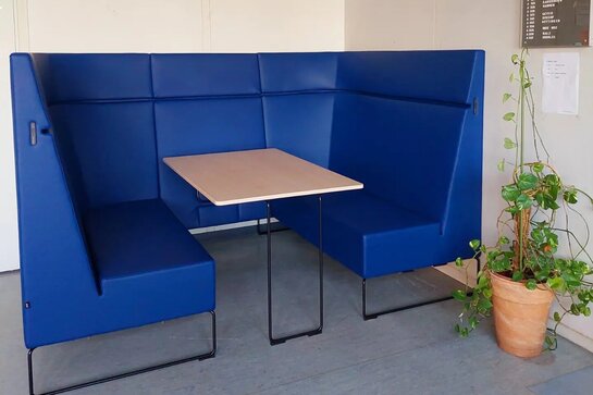 Abgebildet ist eine blaue Lernnische bestehend aus einem Tisch, der von zwei mit hohen Rückenlehnen geschützten Sitzbänken umgeben ist. Daneben steht eine Zimmerpflanze