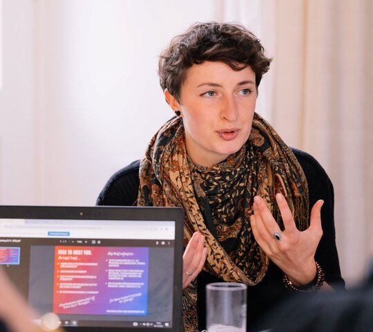 Person in erklärender Pose an einem Tisch, daor Personen mit Blick auf Tablet Bildschirm