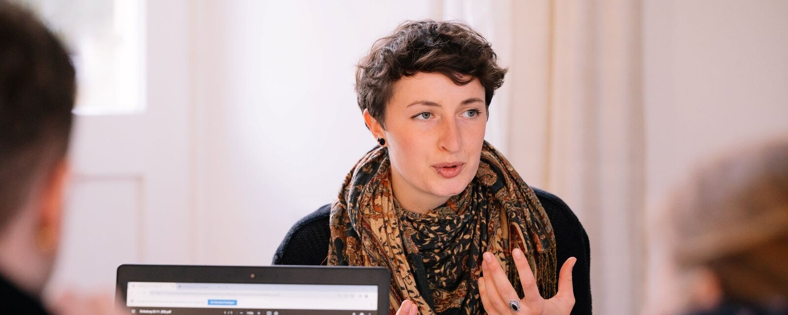 Person in erklärender Pose an einem Tisch, daor Personen mit Blick auf Tablet Bildschirm