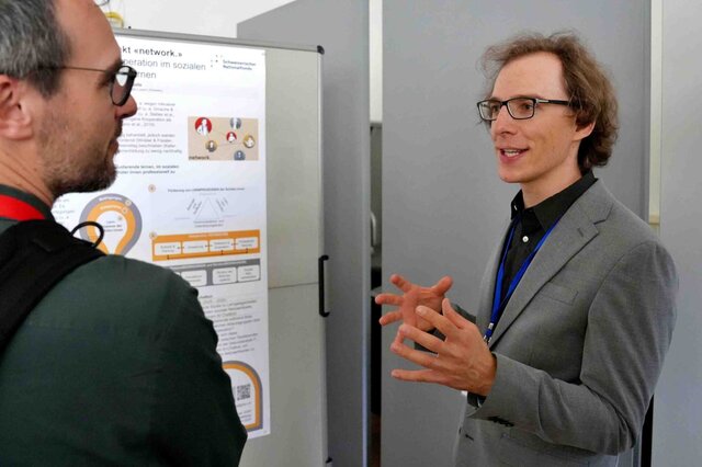 Zwei Männer stehen vor einem Forschungsposter. Der rechte spricht deutlich mit den Händen. Den linken sieht man von schräg hinten.
