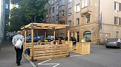 Das Bild zeigt ein sogenanntes Parklet. In diesem sitzen mehrere Menschen. Links im Bild läuft ein Mann auf dem Gehweg; man sieht ihn von hinten. Auf der Straße stehen mehrere Autos.