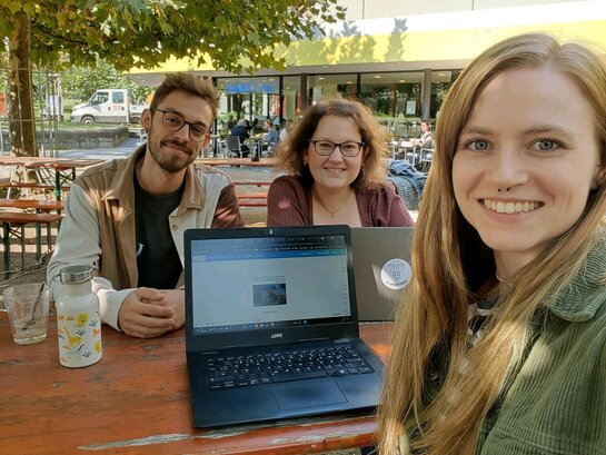 Leo, Chiara und Hannah sitzen an einem Tisch im Freien und lächeln. Auf dem Tisch steht ein Laptop