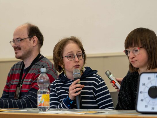 Das Bild zeigt Qualifizierungsteilnehmerin Louisa Kabbe, die in ein Mikrofon spricht. Links von ihr sitzt Bildungsfachkraft Thilo Krahnke, rechts sitzt Qualifizierungsteilnehmerin Susann Bensch.