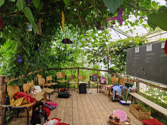 Auf dem Dielenboden einer dschungelartig begrünten Veranda sind Holzstühle auf eine Tafel ausgerichtet, auf der Lebensmittelnamen in Spalten sortiert sind. Auf dem Boden ein Korb mit CO2-Schild sowie weitere Materialien..