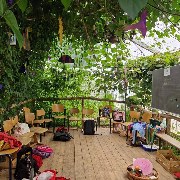 Auf dem Dielenboden einer dschungelartig begrünten Veranda sind Holzstühle auf eine Tafel ausgerichtet, auf der Lebensmittelnamen in Spalten sortiert sind. Auf dem Boden ein Korb mit CO2-Schild sowie weitere Materialien..