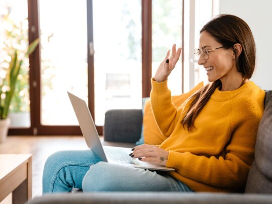 Frau sitzt auf Sofa mit Laptop und winkt in die Kamera
