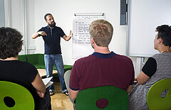 Das Symbolbild zeigt einen jungen Mann an einem Flipchart. Er spricht zu drei Personen, die mit dem Rücken zu dem Betrachter sitzen. Das Bild wurde im sog. Grünen Zimmer der Hochschule aufgenommen. Copyright: PH Heidelberg