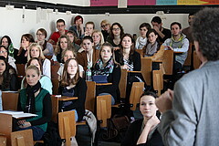 Das Foto zeigt den Rücken eines Lehrenden und Studierende die im Hörsaal sitzen.