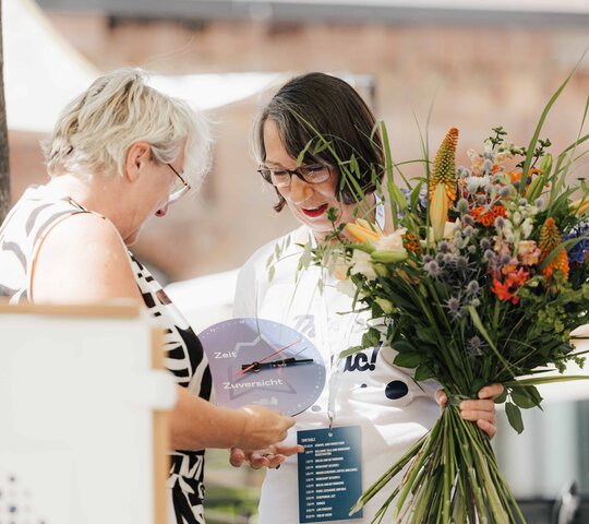 Theresia Bauer, Geschäftsführerin der Baden-Württemberg Stiftung und Wissenschaftsministerin a.D., überreicht Henrike Schön, Leiterin des Akademischen Auslandsamtes der Pädagogischen Hochschule Heidelberg, einen Blumenstrauß sowie eine Uhr.