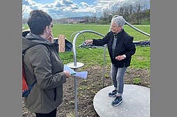 Das Foto zeigt eine ältere Damen auf einem Outdoor-Sportgerät. Eine weitere Person schaut ihr zu.