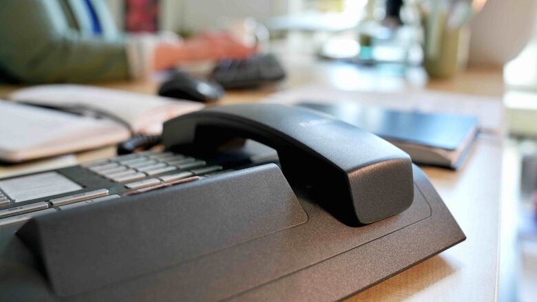 Der Fokus liegt auf einen Festnetztelefon. Im Hintergrund sieht man unscharf eine Person, die an einem Computer arbeitet.