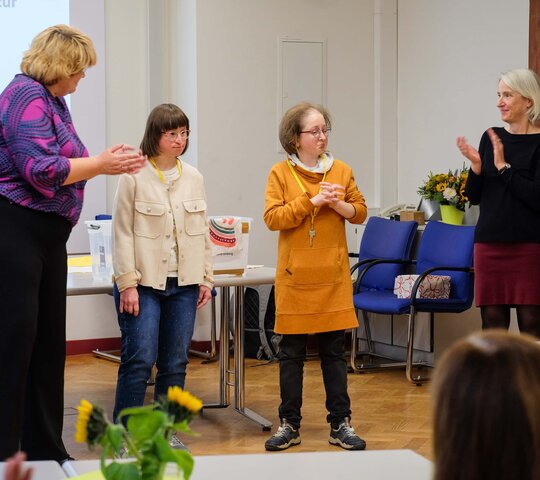 Louisa Kabbe und Susann Bensch sind auf der Feier anlässlich ihrer abgeschlossenn Qualifizierung. Rechts und links stehen die Leiterinnen des AW-ZIB, Prof. Karin Terfloth und Prof. Vera Heyl, und applaudieren.