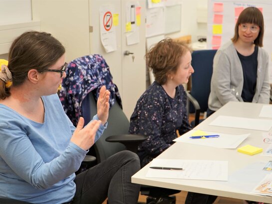 Das Bild zeigt Bildungsfachkraft Anna Neff, die mit den Qualifizierungsteilnehmerinnnen Susann Bensch und Louisa Kabbe in deren Büro spricht. Frau Neff hebt die Hände, um ihr Gesagtes zu unterstreichen.