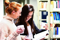 Auf dem Bild sieht man zwei Studentinnen die zusammen in der Bibliothek ein Buch betrachten.