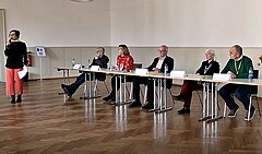 Neben einer stehenden Moderatorin sitzen fünf Personen auf dem Podium und diskutieren.