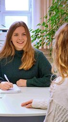Eine Studentin sitzt am Tisch; sie hält einen Stift in der Hand und vor ihr liegen Unterlagen. Sie lächelt eine Frau, die man im rechten Bildrand von hinten sieht, freundlich an.