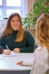 Eine Studentin sitzt am Tisch; sie hält einen Stift in der Hand und vor ihr liegen Unterlagen. Sie lächelt eine Frau, die man im rechten Bildrand von hinten sieht, freundlich an.