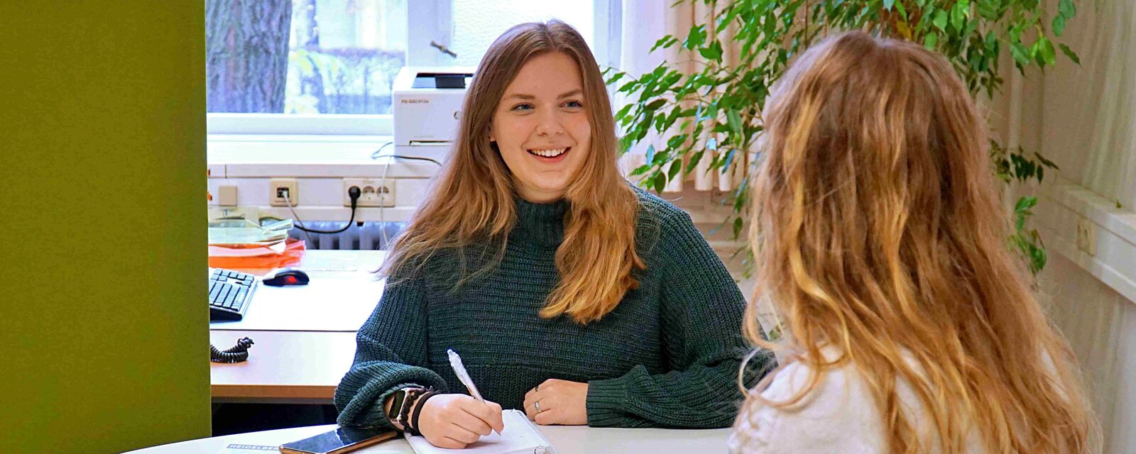 Eine Studentin sitzt am Tisch; sie hält einen Stift in der Hand und vor ihr liegen Unterlagen. Sie lächelt eine Frau, die man im rechten Bildrand von hinten sieht, freundlich an.