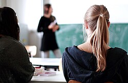 Rücken zweier Studentinnen die eine Lehrende an der Tafel anschauen.