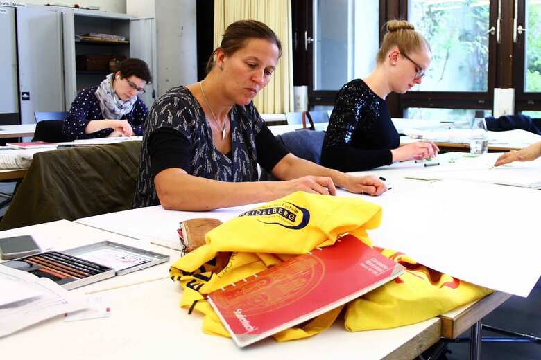 Drei Student:innen arbeiten konzentriert im Seminarraum. Auf dem Tisch liegt - gut sichtbar - eine Tasche mit dem Logo der PHHD sowie eine Broschüre mit der Logo der Universität Heidelberg.