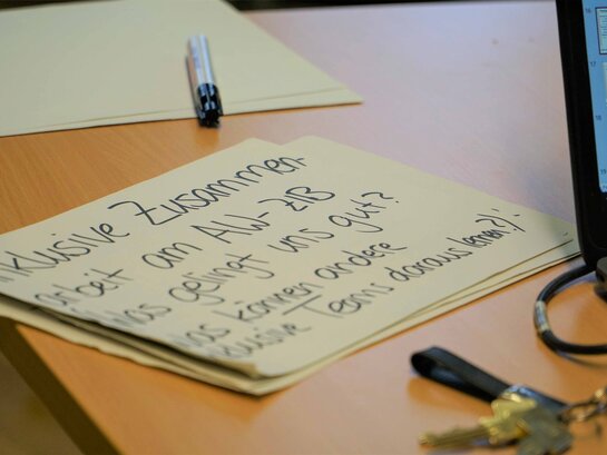 Das Bild zeigt eine gelbe Karteikarte, die auf einem Tisch liegt. Darauf steht in schwarzer Schrift geschrieben: Inklusive Zusammenarbeit am AW-ZIB. Was gelingt uns gut? Was können andere inklusive Teams daraus lernen?